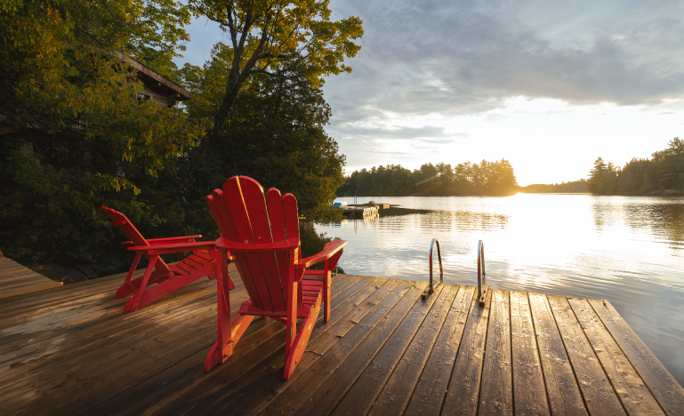 Scenic view of a wooden dock with red Adirondack chairs overlooking a calm lake at sunset.