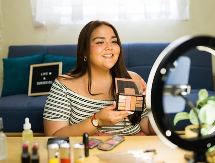a young woman is seated at vanity table holding an open palette of eye shadow and staring into a camera with a ring light around it as she talks about the product