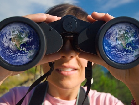 women standing outside in nature holds binoculars up to her eyes and we see reflection of globe in both lenses