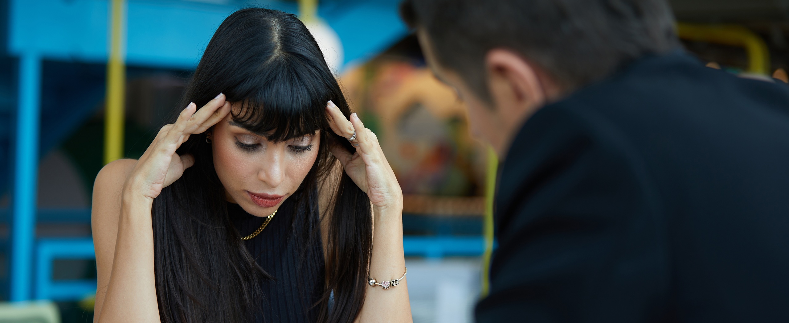 young woman seated across from older, suited professional at table, head down, massaging temples, looking stressed