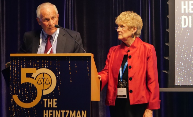 Elderly man speaking at a podium with a '50 The Heintzman' sign, while an elderly woman in a red jacket stands beside him.