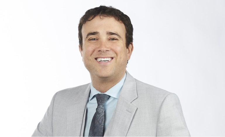 Smiling man in a light gray suit, blue shirt, and patterned tie against a white background.