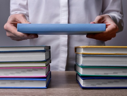 hands clasping a book, positioning it in a bridge-life-fashion over two side-by-side stacks of books on a table 