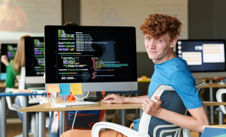 Young male programmer in a blue shirt sitting at a desk with a computer screen displaying code in a classroom setting.