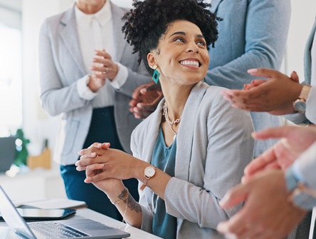 a women seated in front of computer at desk is surprised and delighted to be surrounded by coworkers who are applauding her work