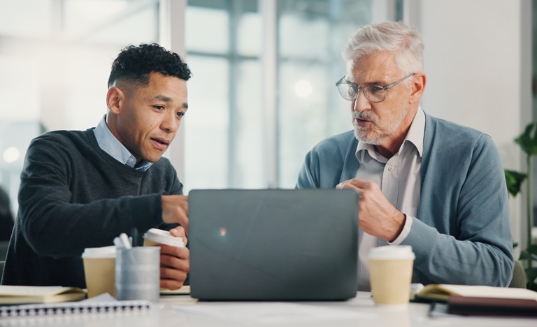 A young male lawyer sits next to an older male client at a table explaining information that appears on the laptop screen in front of them