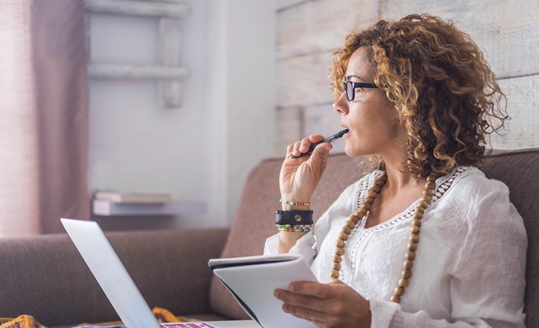 A woman sits on a couch with her laptop on her lap, a notepad in her, and his the tip of the pencil in her mouth as she gaze into the distance in deep thought