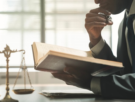 a man seated at a desk with a hand beneath his chin studies a legal book that is open before him next to table-top scales of justice