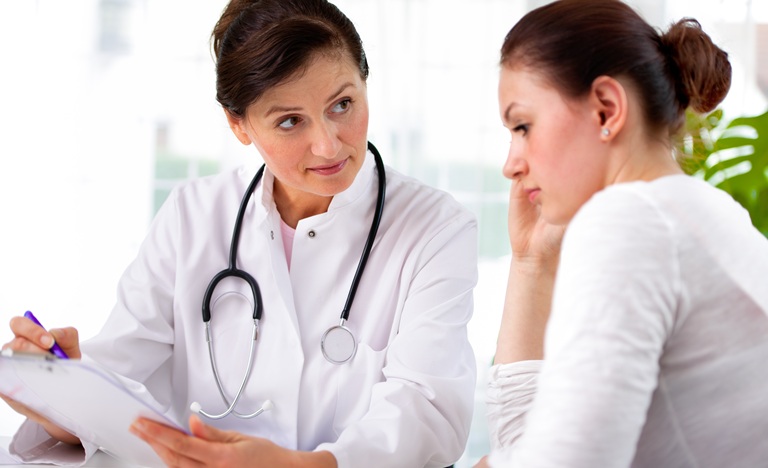 Female doctor in white coat seated at table with clipboard in hand explaining to female patient, seated beside her, what is on the chart