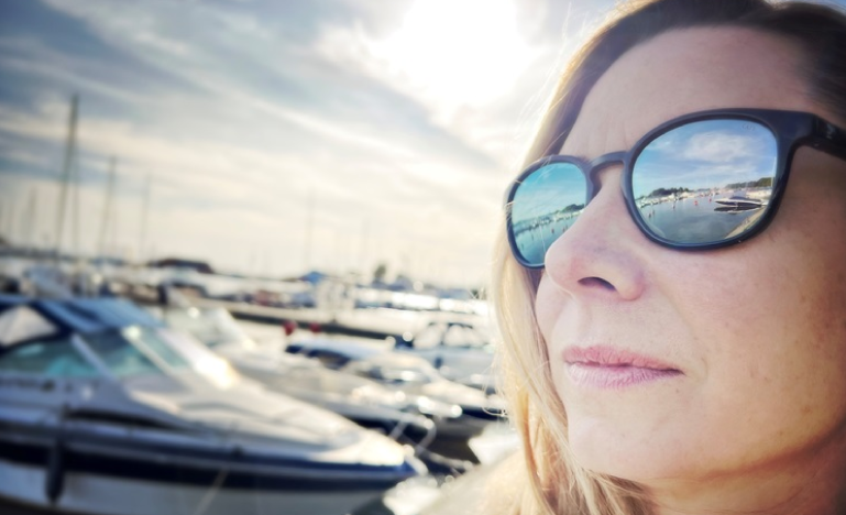 Gros plan d'une femme portant des lunettes de soleil reflétant une marina avec des bateaux sous un ciel ensoleillé."