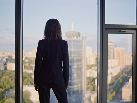 Woman in a business suit stands with her back to the camera, looking out the floor-to-ceiling window of an office at the skyscrapers and buildings and open sky around her