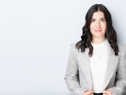 Photo of OBA President Katy Commisso standing in a grey jacket and white t-shirt, smiling, with hands at waist, against like grey background