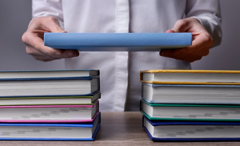 hands clasping a book, positioning it in a bridge-life-fashion over two side-by-side stacks of books on a table 