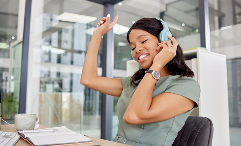 Woman sitting at office-desk with headphones on, smiling, with one hand raised snapping to the music