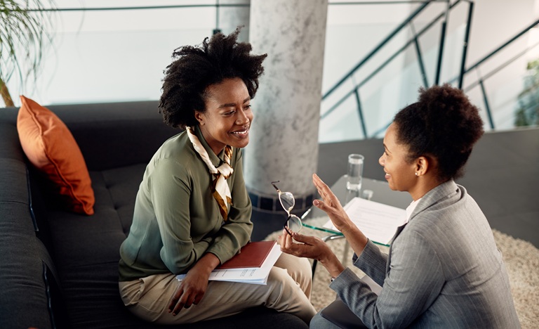 two women, one young, one middle-aged, sit across from each other in engaged conversation, while the younger woman takes notes and the older leans in with advice