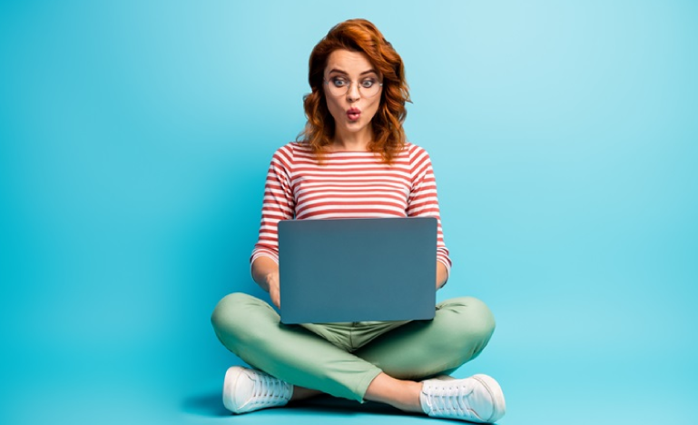 Surprised woman with red hair and glasses sitting cross-legged with a laptop against a blue background.