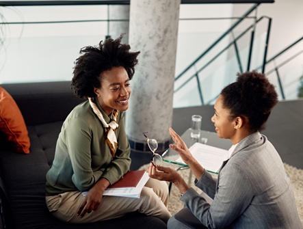 two women, one young, one middle-aged, sit across from each other in engaged conversation, while the younger woman takes notes and the older leans in with advice